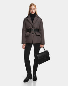 A woman stands against a white background wearing the Rudsak PAOLA - 8124547 Grey down blazer, paired with a black turtleneck, black pants, knee-high black boots, and holding a black handbag at her side.