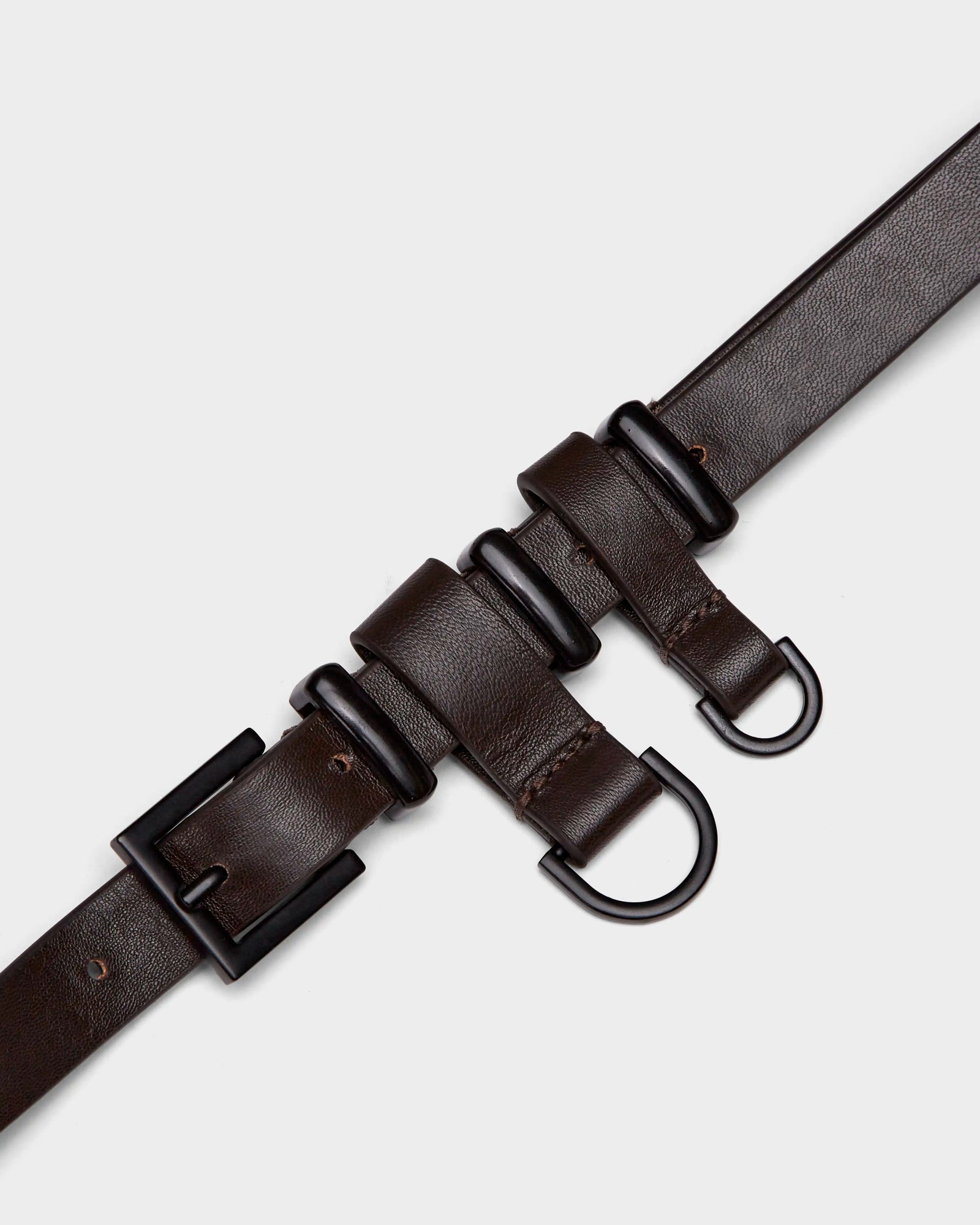 The PREYA - 8324508 Espresso belt by Rudsak is a dark brown leather accessory with a black rectangular buckle, two black D-rings, and three leather loops, offering a modern utilitarian look displayed against a light grey background.