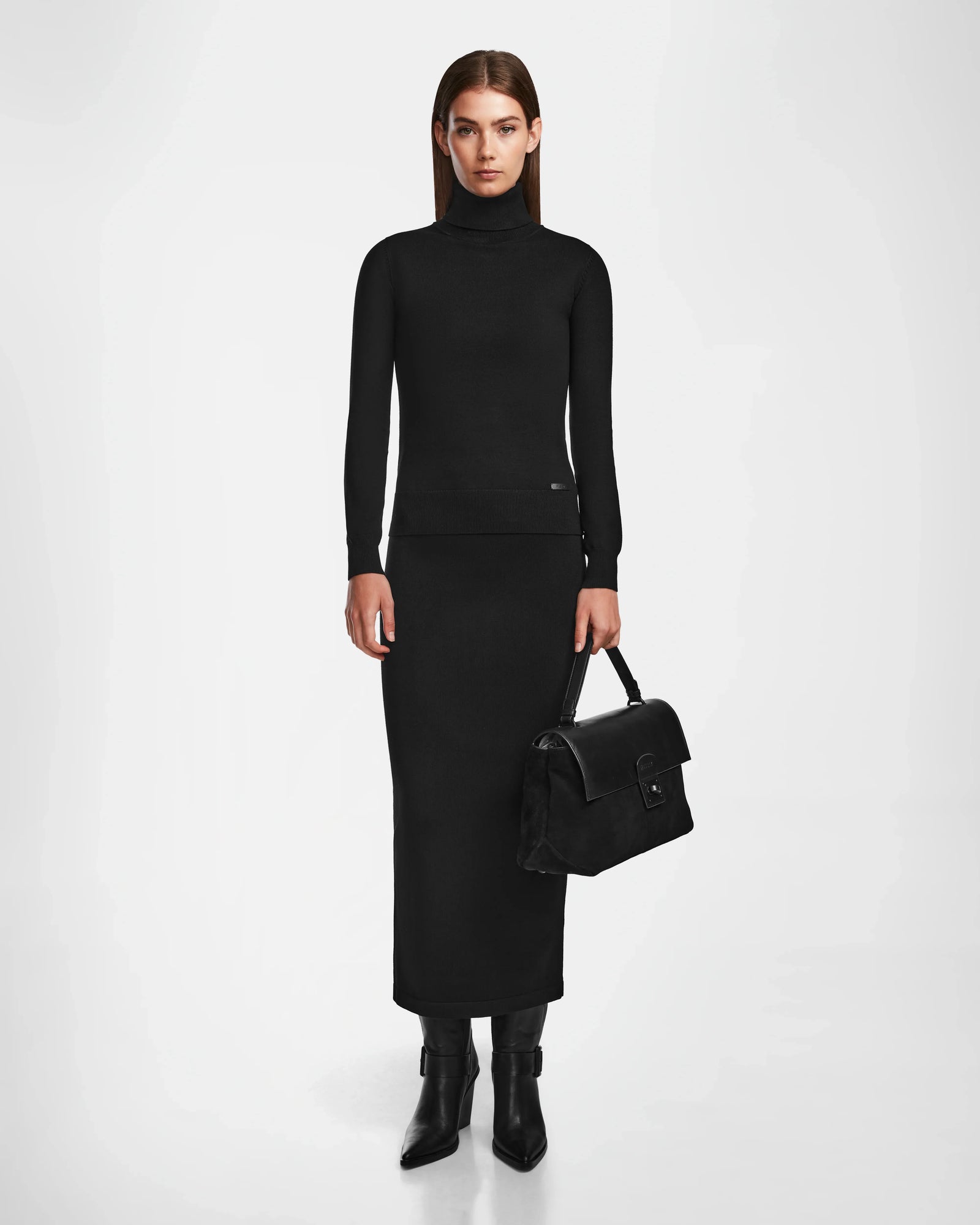 A woman stands against a white background wearing a fitted black turtleneck, long matching skirt, black pointed boots, and holding the Rudsak ESMA - 8324519 Black suede and leather handbag with removable shoulder strap by her side.