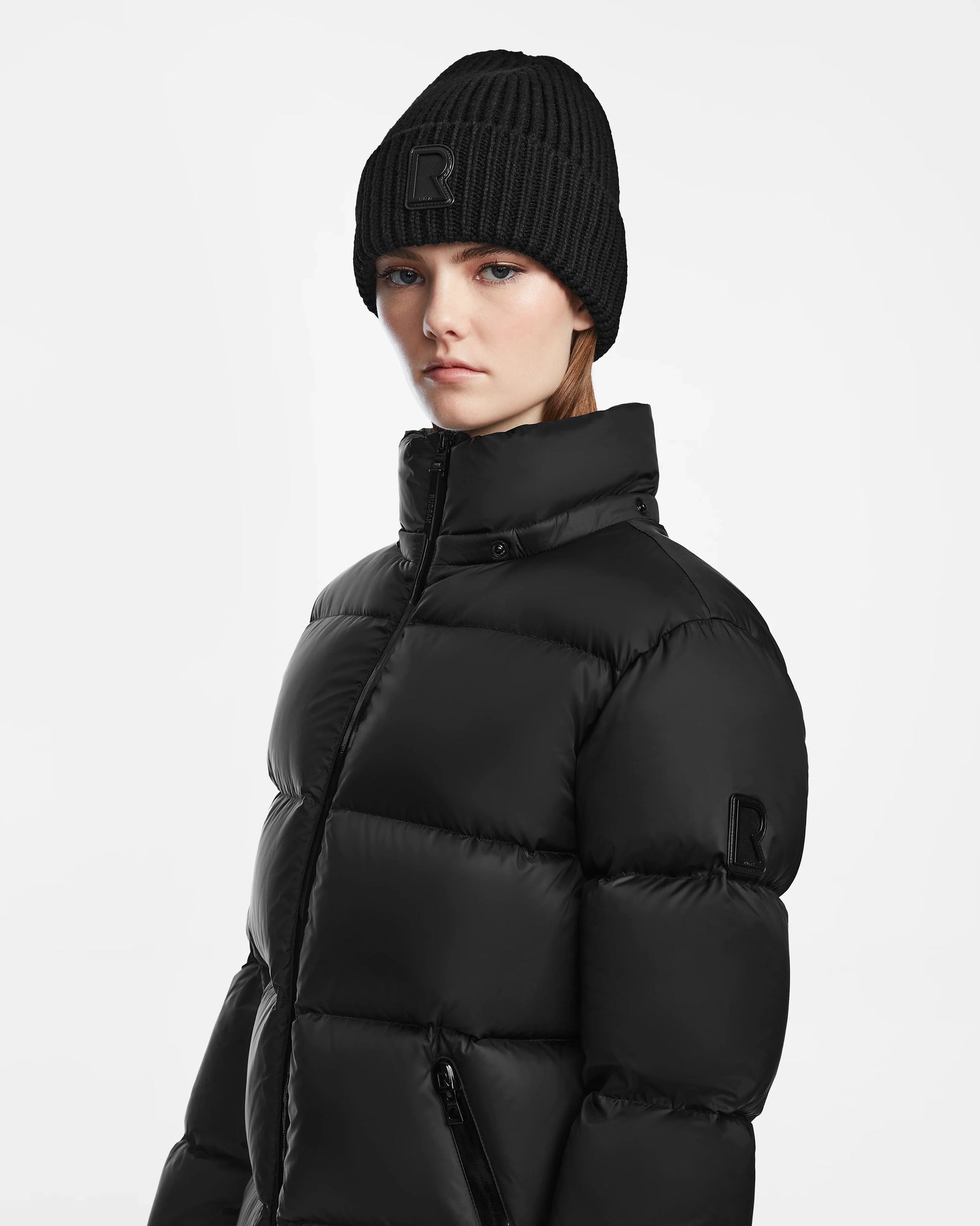 A woman wearing the Rudsak RITA M - L124537 Black puffer jacket and a black knit beanie stands against a white background, looking straight ahead with a neutral expression.