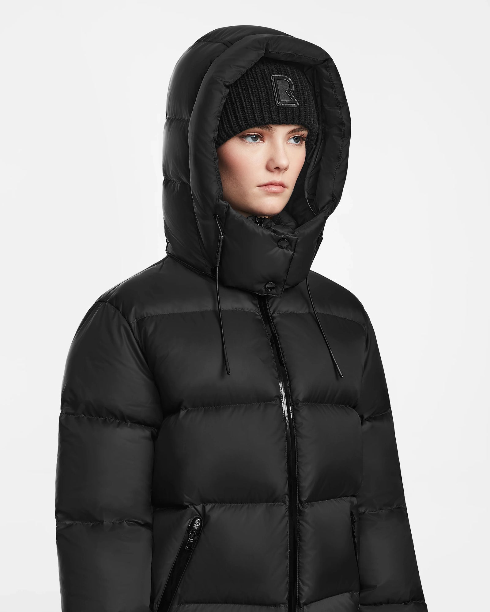 A person wears the Rudsak RITA M - L124537 Black women’s hooded down puffer with RDS-certified down, the hood up over a black beanie, looking forward with a neutral expression against a plain white background.