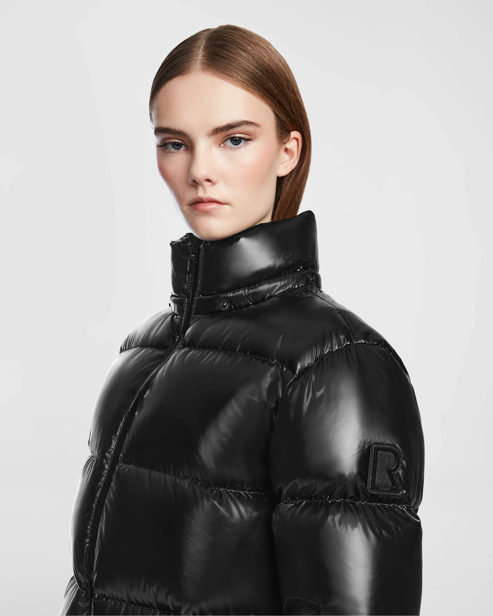 A woman with straight brown hair and natural makeup wears the Rudsak RITA S - L124520 Black, a shiny, weather-resistant puffer jacket with a high collar and embossed R patch on the sleeve, posing against a light gray backdrop.