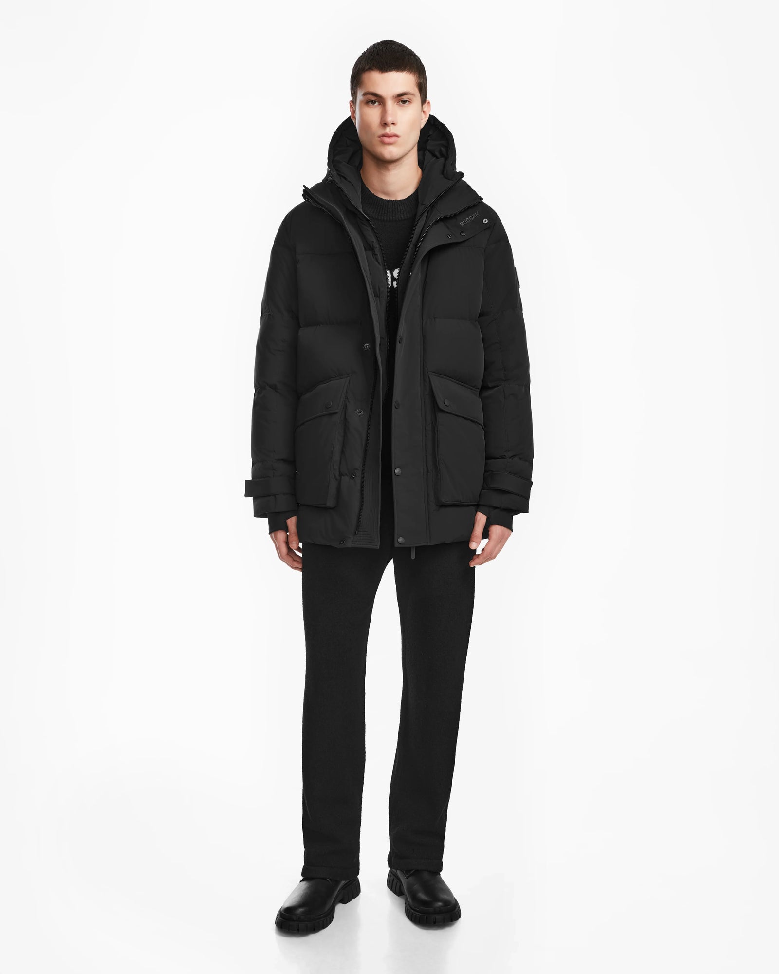 A person faces forward against a white background, wearing the Rudsak ROKO - 6124510 Black men’s quilted down parka with hood, large front pockets, and RDS-certified insulation, paired with black pants and shoes.