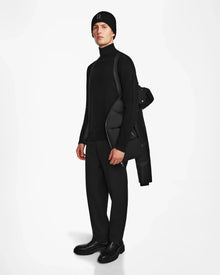 A person models Rudsaks RONNY M - M124513 Black winter puffer, crafted from recycled materials, draped over one shoulder while dressed in all black, standing against a plain white background.