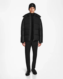 A person wears the Rudsak RONNY M - M124513 Black puffer jacket, paired with black pants, boots, and a beanie, standing against a plain white background facing forward with arms at their sides.