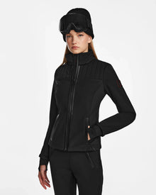 A woman wears the Rudsak ROSALIE - 8124784 Black technical ski jacket with RECCO rescue technology, black pants, a knit beanie, and ski goggles, standing against a white background with her hands in her jacket pockets.