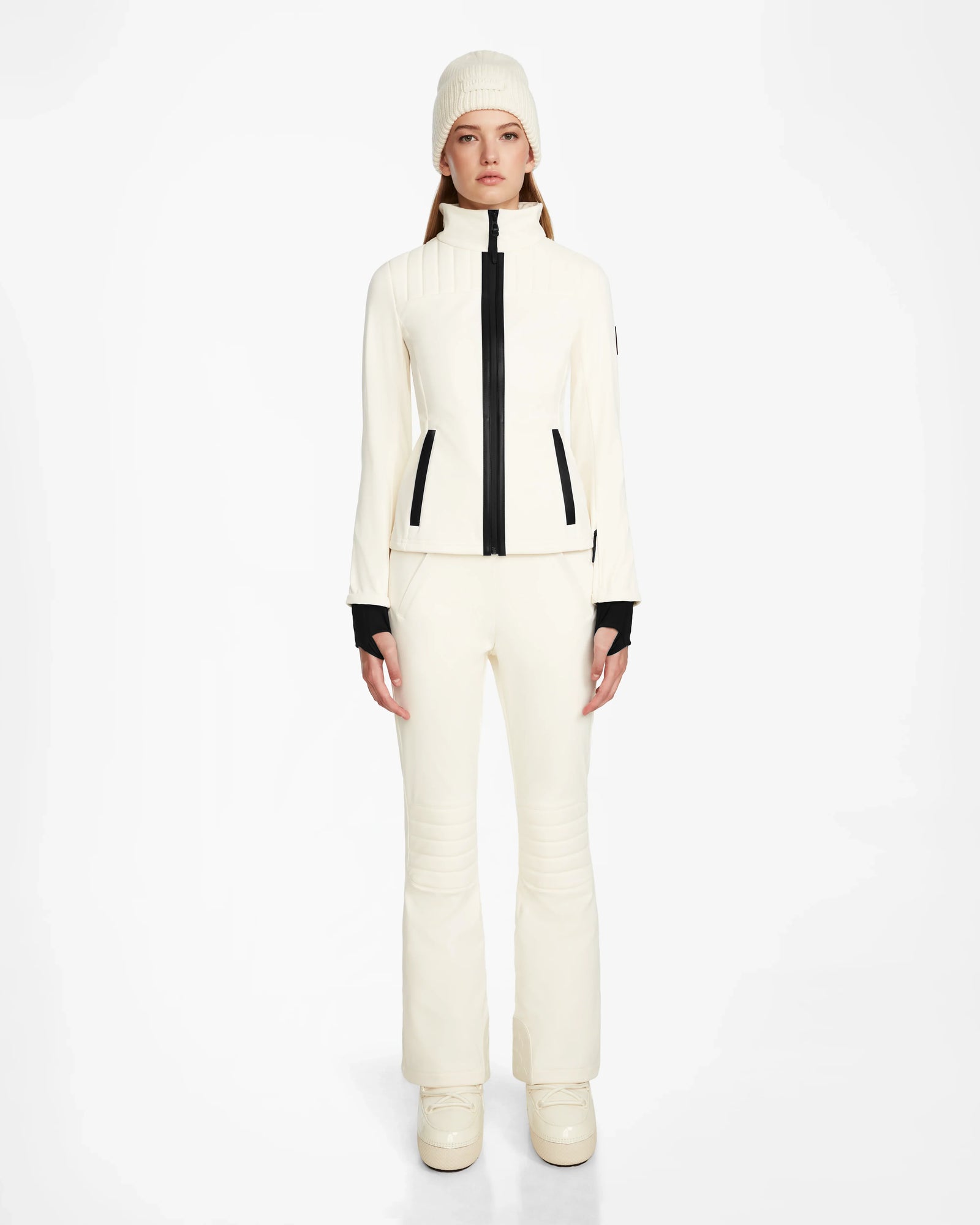 A woman faces forward in the ROSALIE - 8124784 Iceland women's fitted ski jacket by Rudsak with black accents, matching pants, a knit beanie, and platform boots. The high-performance jacket features RECCO rescue technology for extra safety.