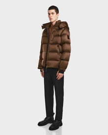 A young man stands facing forward against a plain background, wearing the Rudsak VINCE S - 6124560 Antique Bronze mens quilted down puffer, dark trousers, black dress shoes, and a brown turtleneck sweater.