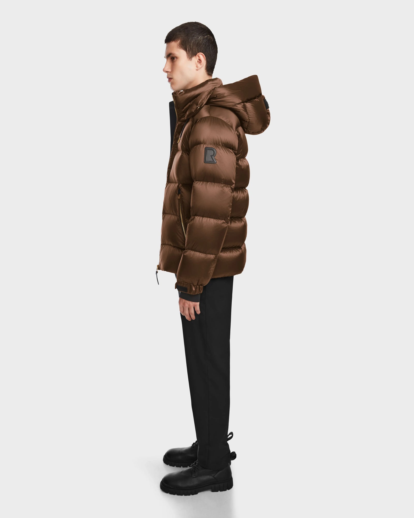 A person stands in profile wearing the Rudsak VINCE S - 6124560 Antique Bronze men’s quilted down puffer jacket with a hood, paired with black pants and boots, against a plain light background.