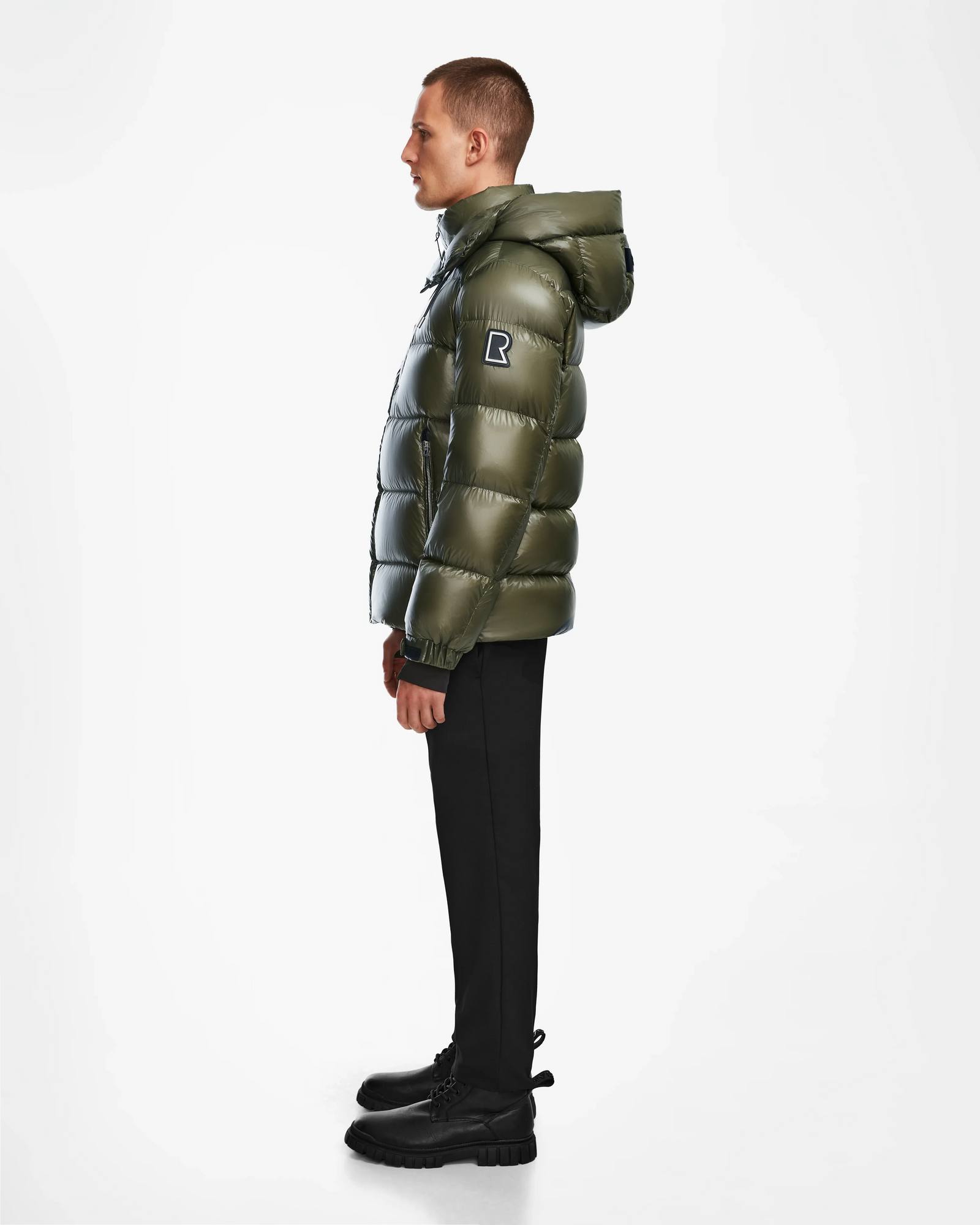 A person stands in profile wearing the Rudsak VINCE S - 6124560 Loden Green mens down puffer jacket with a hood, black pants, and black boots against a plain white background.