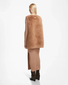 A person faces away wearing the Rudsak YOLANDA - 8124790 Camel faux fur vest over a ribbed tan knit dress with a side slit and dark brown boots. Their straight blond hair is tucked behind one ear. Perfect for fall and winter. White background.