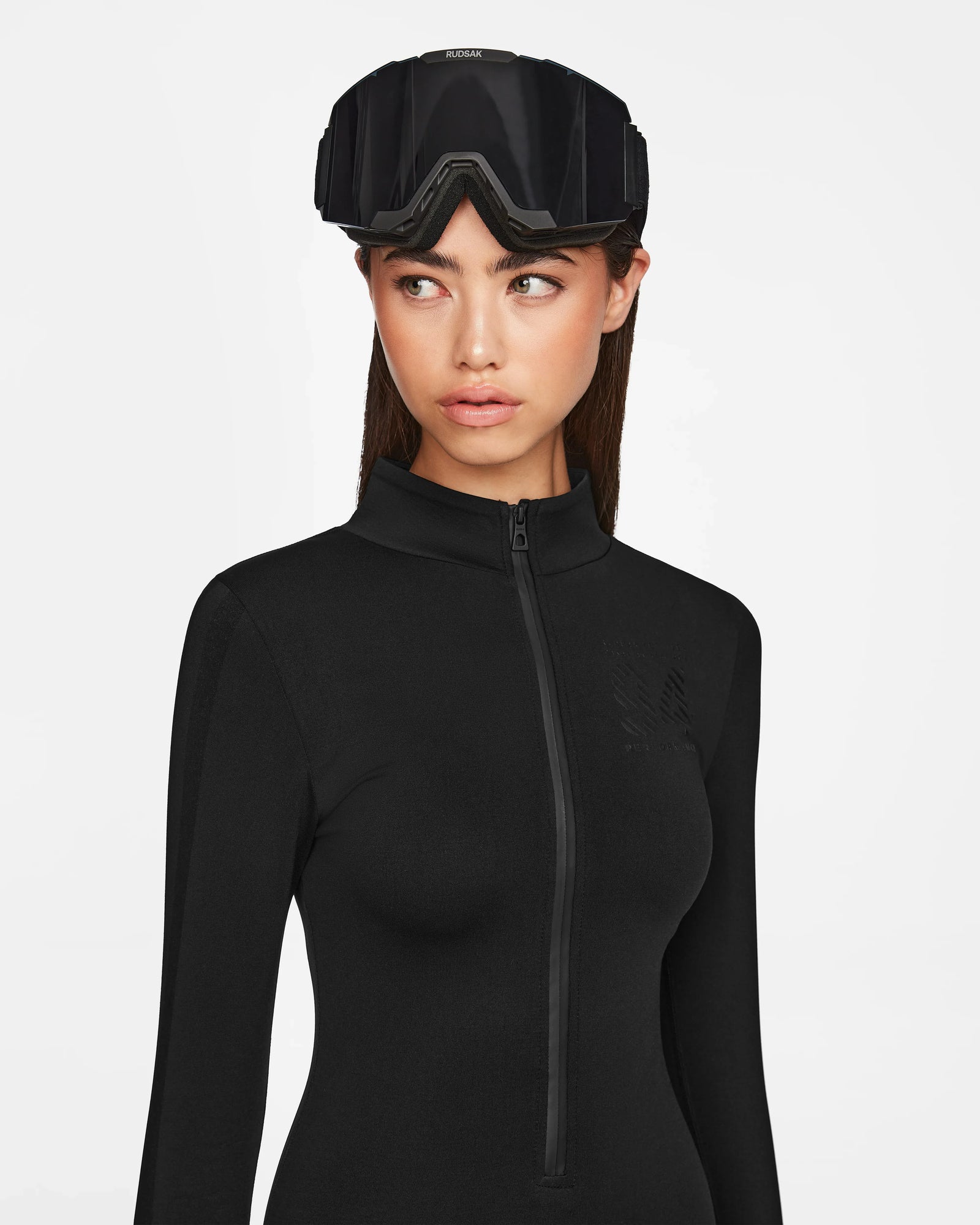 A woman wearing the Rudsak ZADIE - 8524609 Black long sleeve bodysuit and large black ski goggles on her head looks to the side against a plain white background.