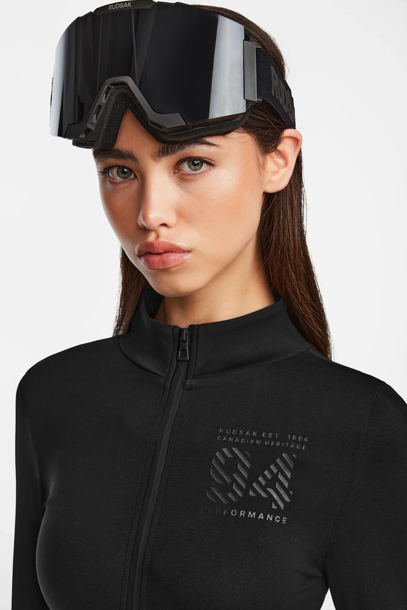 A woman with long brown hair wears black ski goggles on her forehead and the Rudsak ZADIE - 8524609 Black zip-up jacket, featuring white text and a logo on the chest, against a plain white background.
