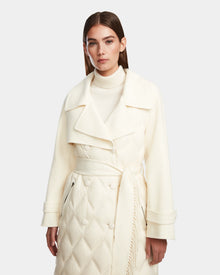 A woman with straight brown hair models the Rudsak ZOE - 8124711 Iceland cream long trench coat, featuring a wide collar, double-breasted buttons, and waist tie, paired with a matching turtleneck against a plain background.