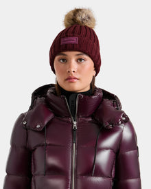 A person wearing the Rudsak ANISA - L423500 Wine puffer jacket and a matching maroon women's knit hat with faux fur pom-pom faces forward against a plain background, highlighting chic winter essentials.