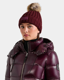 Wearing the Rudsak ANISA - L423500 Wine women’s knit pom pom hat with faux fur and a shiny, dark purple puffer jacket, she looks forward with a neutral expression—an ideal pick for your winter wardrobe.