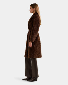 A woman in profile wears the Rudsak ANNABELLE FSU - L125512 Espresso long faux suede trench coat with a waist tie, paired with dark brown pants and brown wedge boots, set against a plain light background.