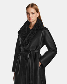 A woman with straight, light brown hair wears the Rudsak ANNABELLE - L124544 Black vegan leather coat with a large textured black fur collar and belt, standing against a plain light background.