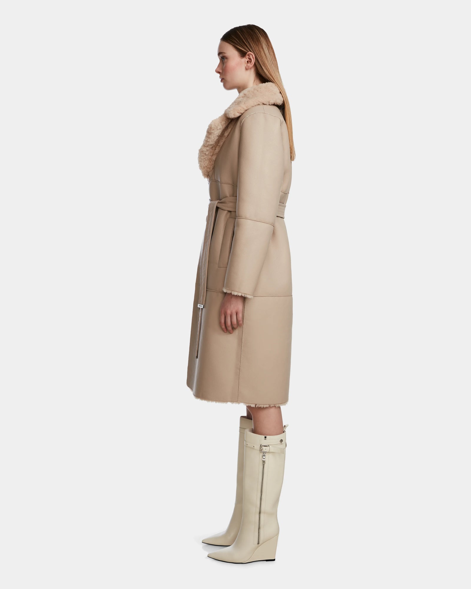 A woman in profile wears the Rudsak ANNABELLE - L124544 Sand women's reversible teddy coat with a shearling collar and belt, paired with knee-high off-white boots. She has straight light brown hair and stands against a neutral background.