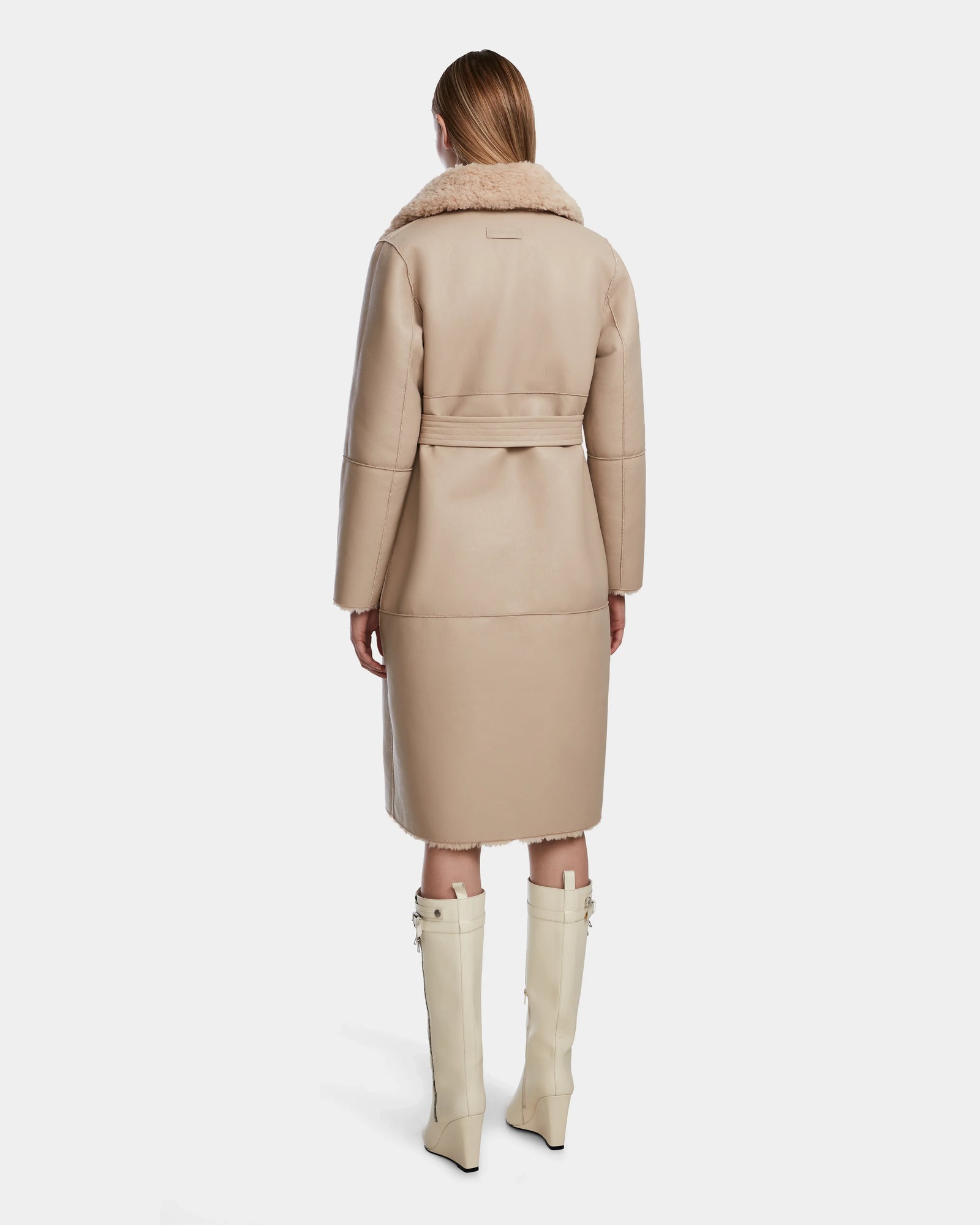 A woman faces away, wearing the Rudsak ANNABELLE - L124544 Sand knee-length shearling coat with a wide fur collar and matching belt, paired with cream knee-high boots featuring decorative straps.