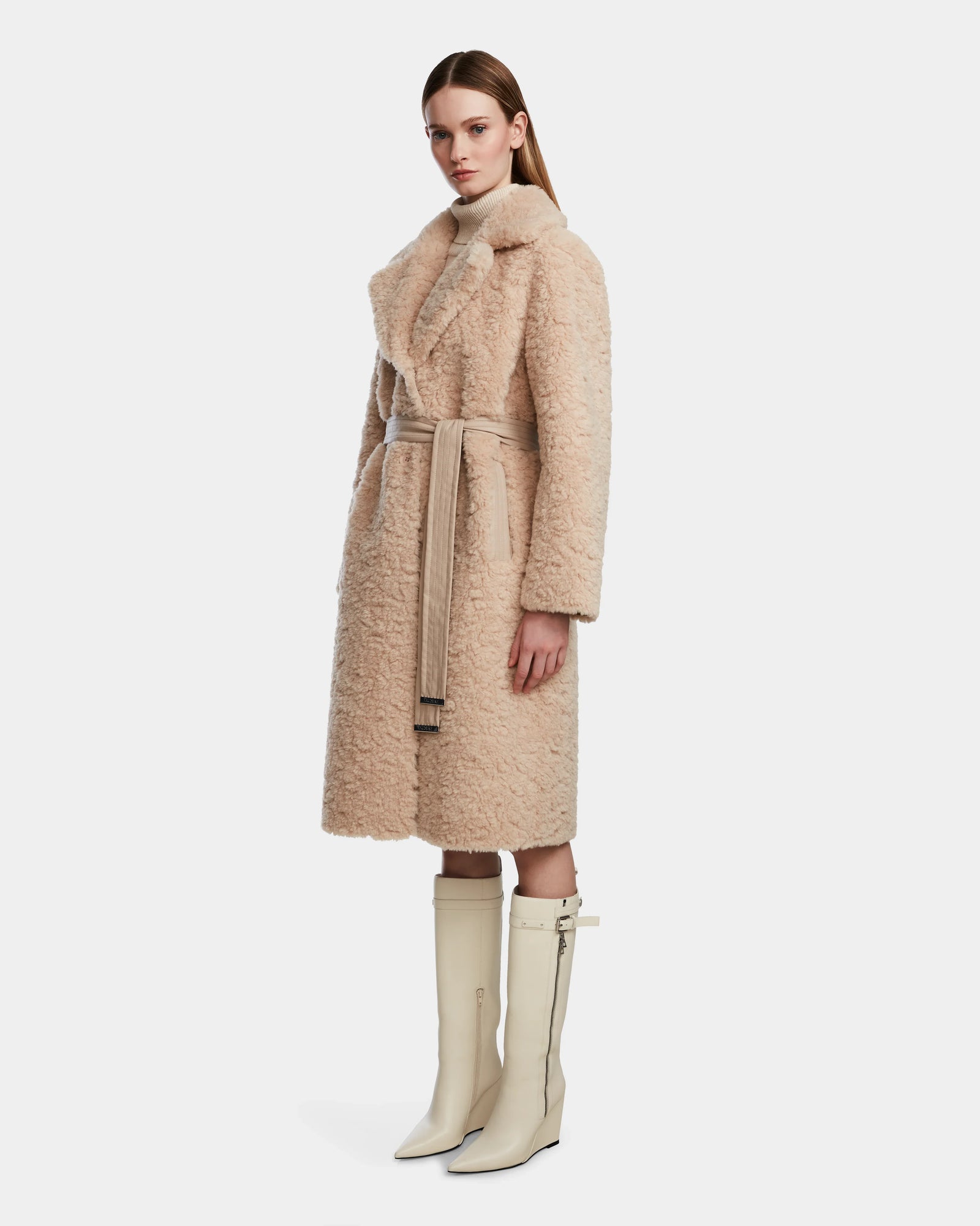 A woman wearing the Rudsak ANNABELLE - L124544 Sand belted faux shearling coat and cream knee-high heeled boots stands against a plain white background, looking forward with a neutral expression.