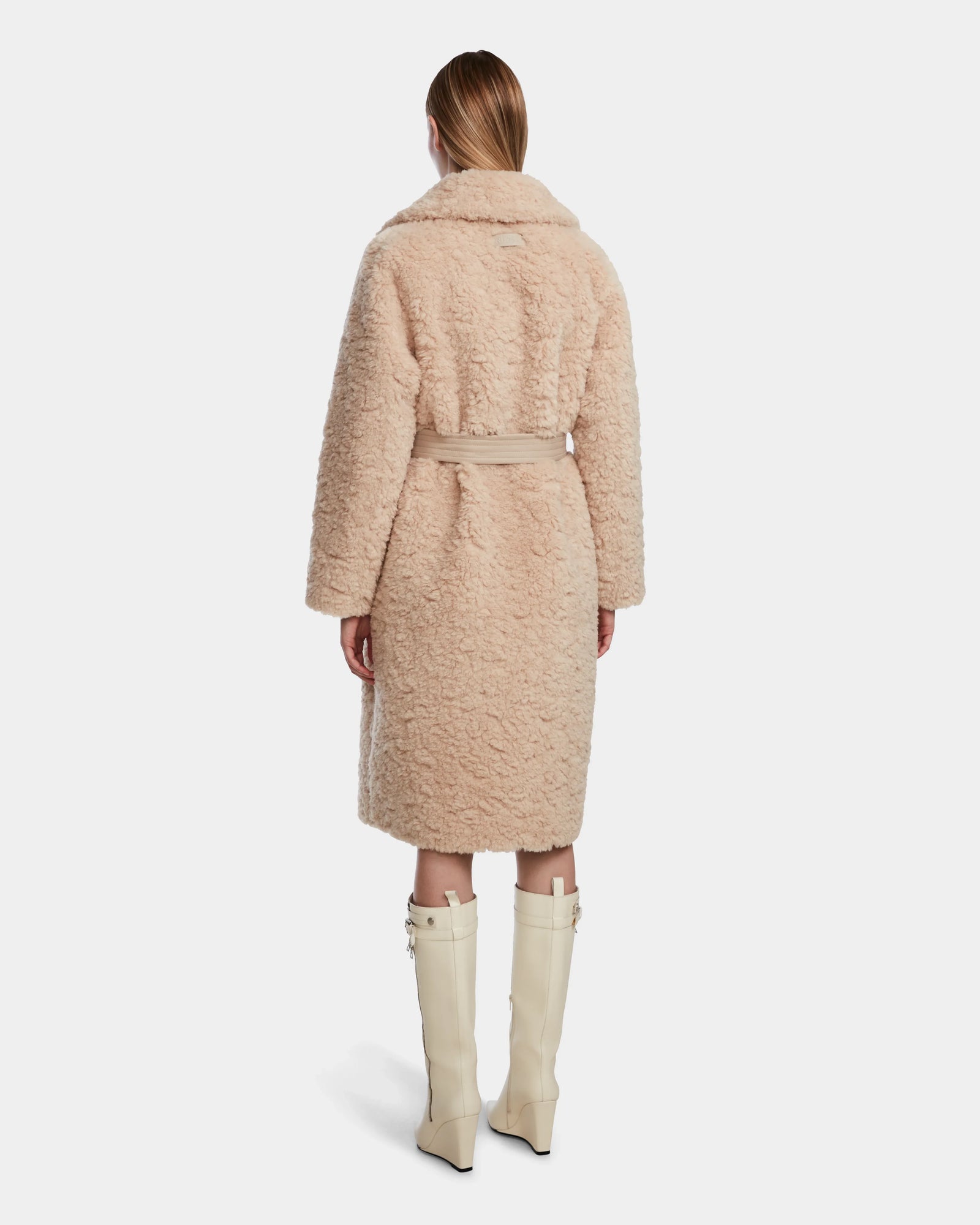 A woman stands facing away, wearing the Rudsak ANNABELLE - L124544 Sand reversible teddy coat and cream knee-high heeled boots. Her straight hair is down, posed against a plain white background.