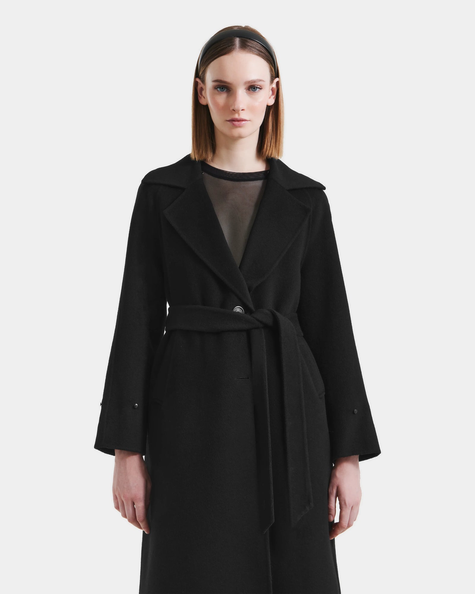 A woman with straight, shoulder-length hair models the Rudsak ARTIS - L125575 Black women’s wool coat with a belted waist over a sheer black top, standing against a plain white background and facing forward neutrally.