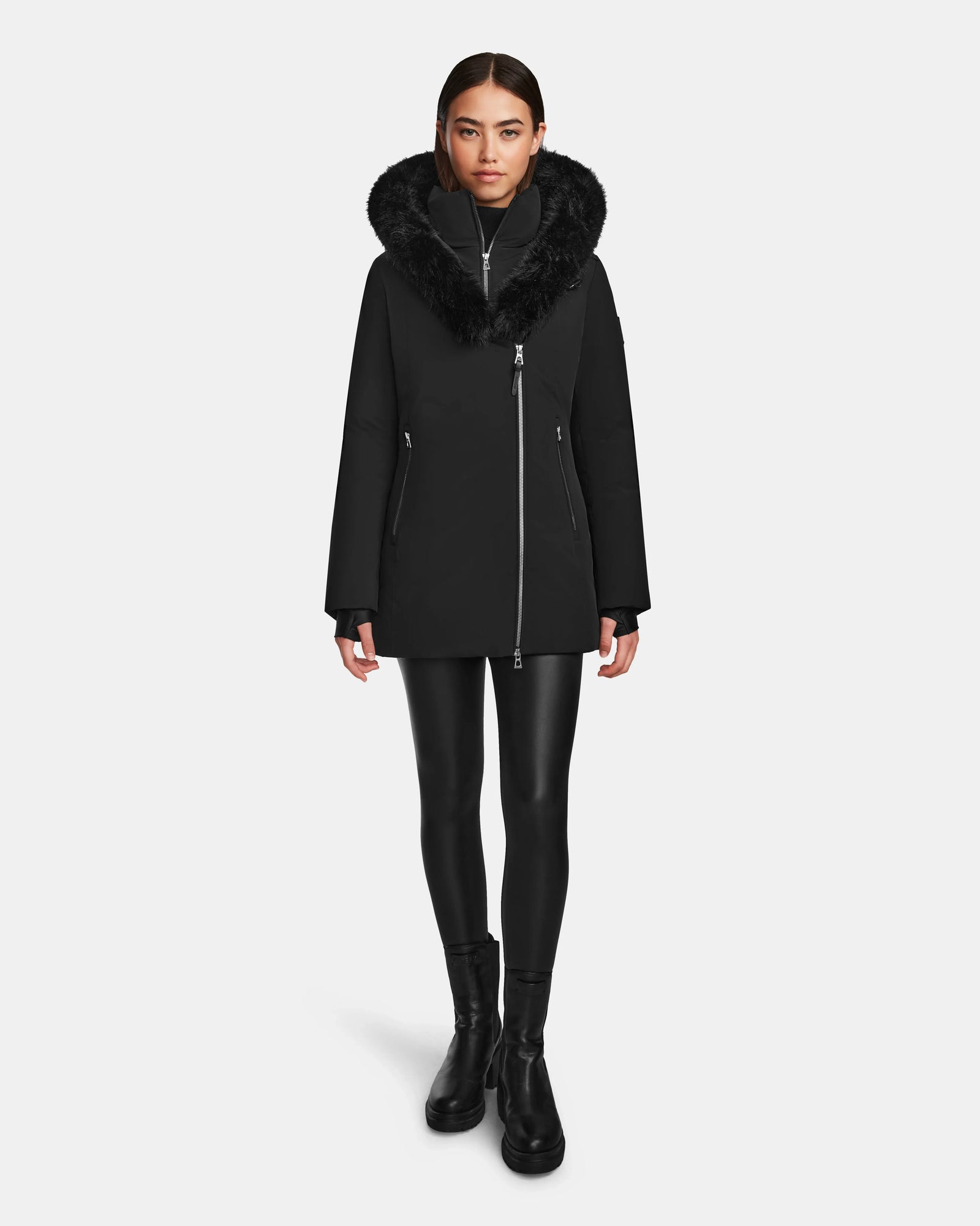 A woman stands facing forward wearing the Rudsak AURORE - L123561 Black/Black women’s short fitted down parka with faux fur trim, paired with black leggings and black ankle boots, against a plain white background.