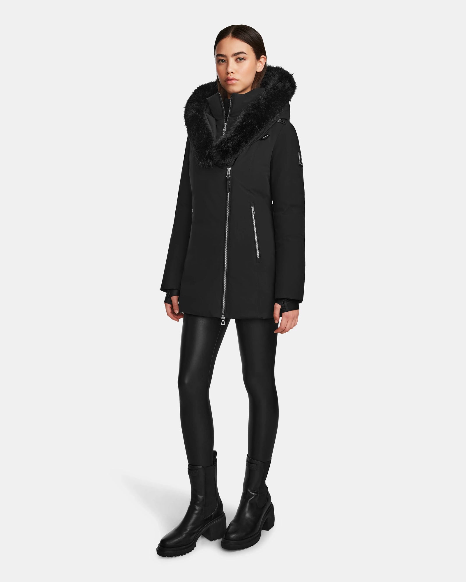 A woman stands against a plain white background wearing the Rudsak AURORE - L123561 Black/Black women's short fitted down parka with a fur-trimmed hood, paired with black leggings and black boots.