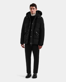 A man faces forward wearing the Rudsak BRADY - M125510 Black men’s quilted down parka with removable hood, paired with black pants, black shoes, and a white turtleneck against a plain light gray background.