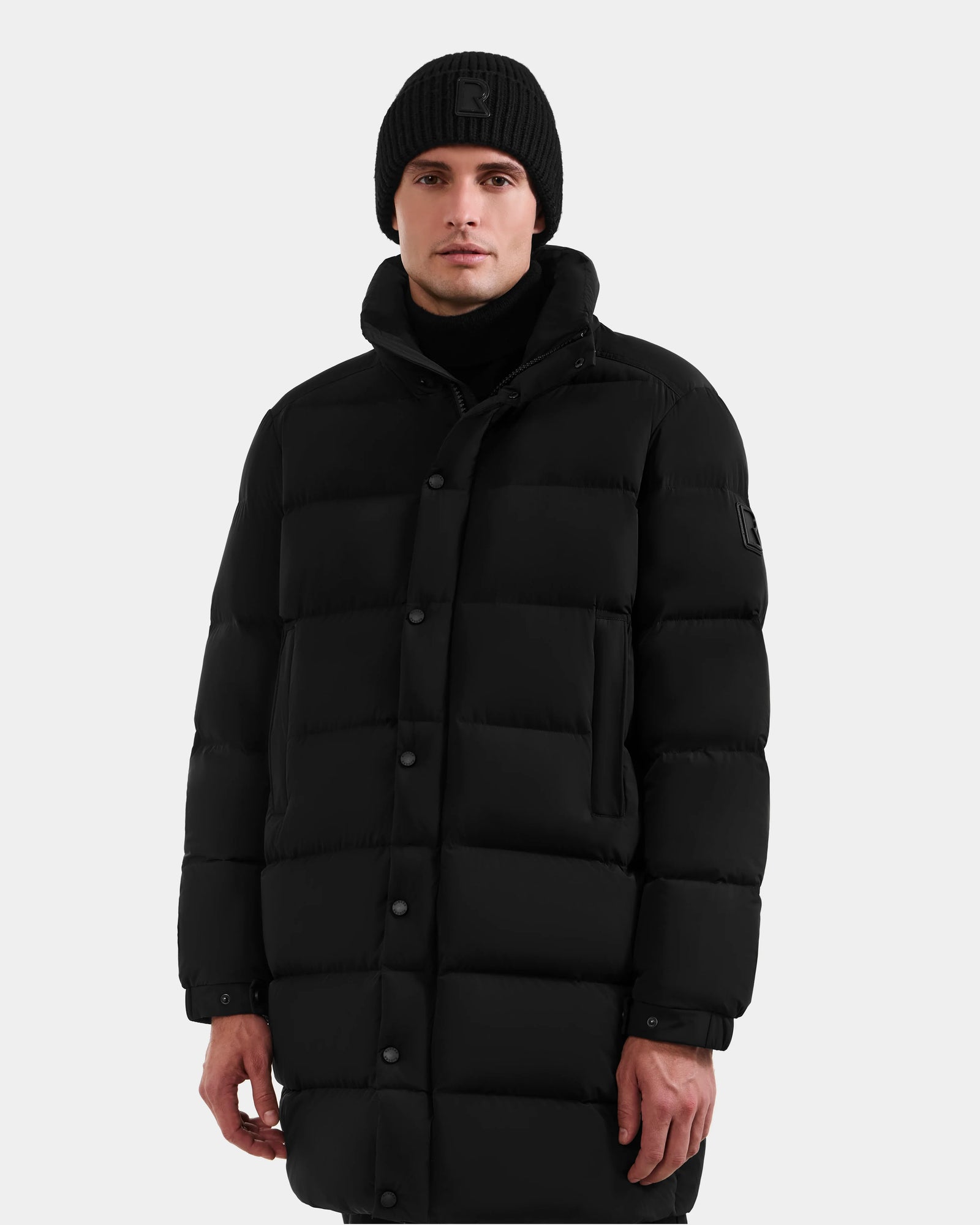 A man wearing the Rudsak BRAYDEN M-M - M125535 Black down puffer coat, a black knit beanie, and a black turtleneck stands against a plain light gray background, looking directly at the camera.