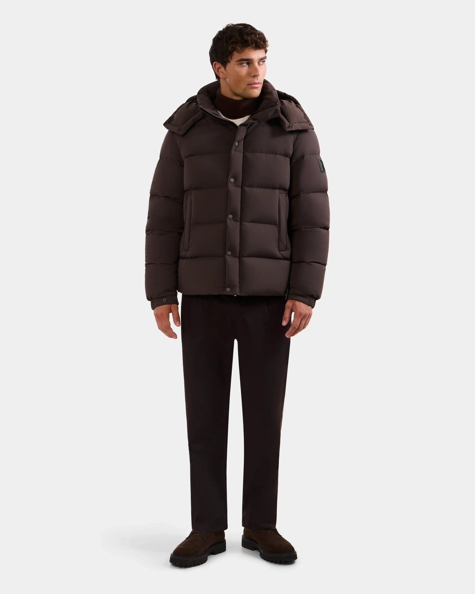 A man stands against a white background wearing the Rudsak BRAYDEN M-S - M125524 Espresso men’s down puffer jacket with hood, paired with dark trousers and brown shoes. He looks slightly to the side, arms relaxed at his sides.