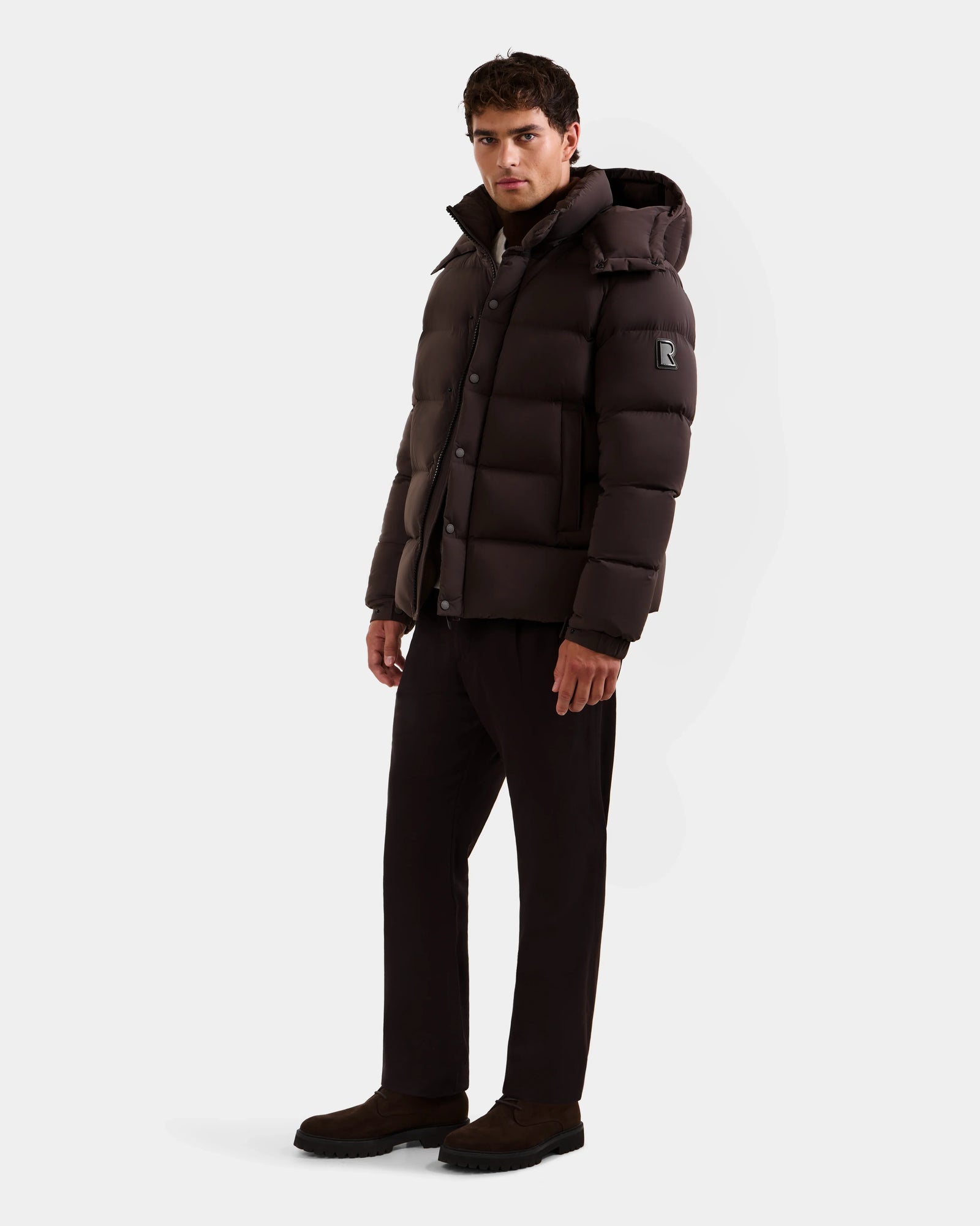 A man stands against a plain white background wearing the Rudsak BRAYDEN M-S - M125524 Espresso men’s down puffer jacket with a hood, dark trousers, and shoes. The winter jacket features an upper sleeve patch with the letter R.