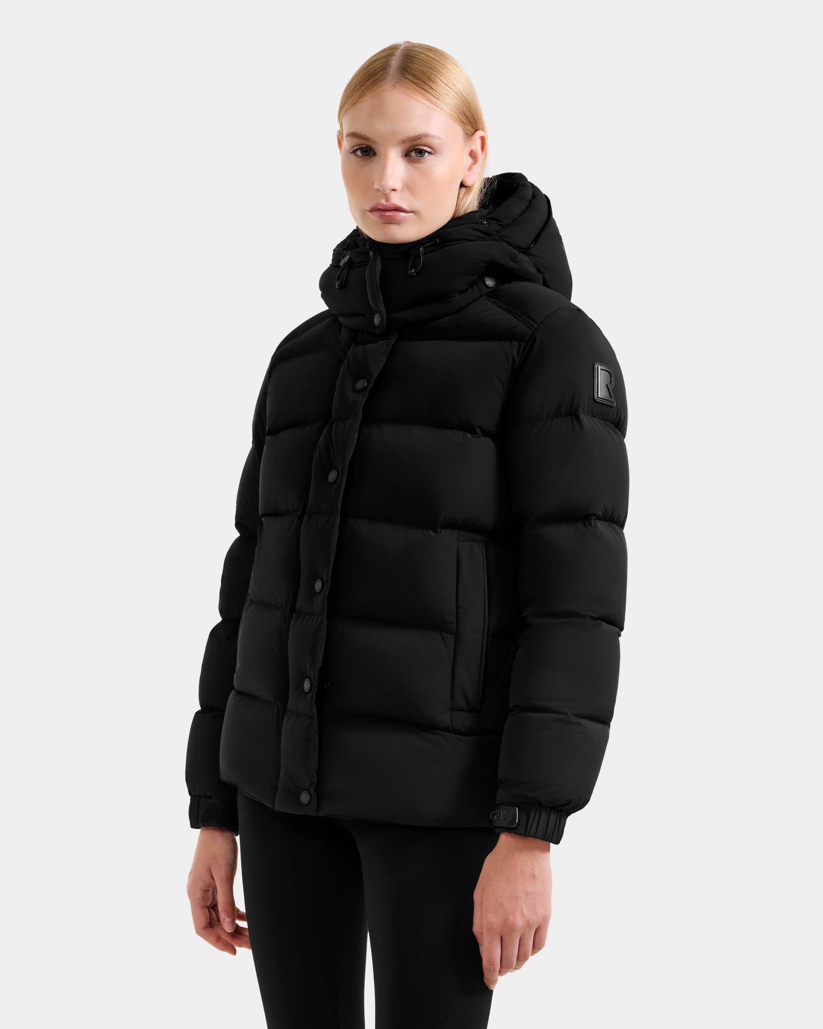 A woman with straight blonde hair wears the Rudsak BREE M - L125582 Black women’s down puffer jacket with large horizontal quilting and a removable hood, paired with black pants, standing against a plain light gray background.