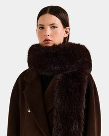 A woman with straight brown hair models the Rudsak CARMY - L425534 Espresso, a plush women’s faux-fur scarf, wrapped around her neck and paired with a dark brown coat against a plain light background.