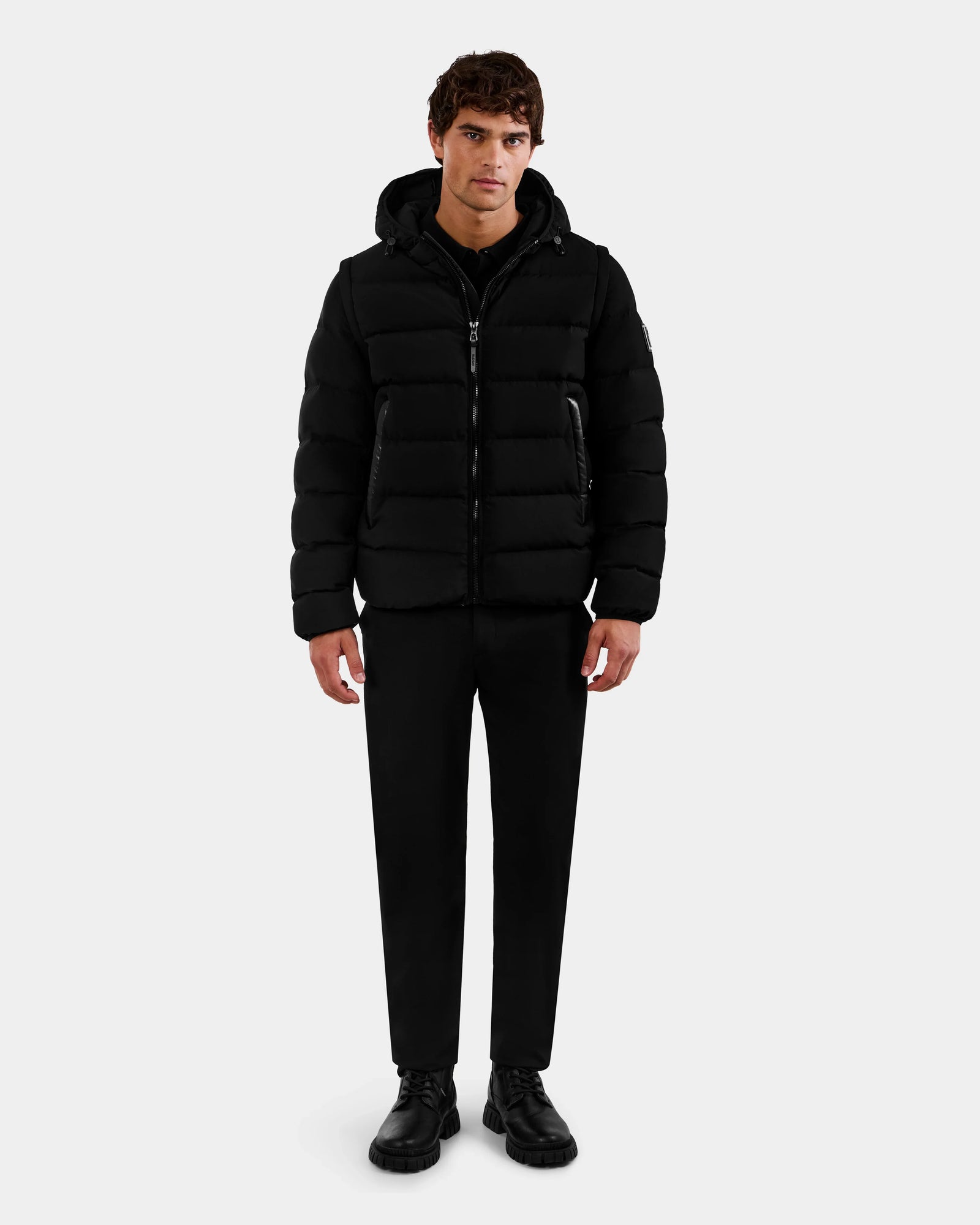 A man stands facing forward wearing the Rudsak DEMIAN M-R - M123504 Black men's down puffer jacket, black pants, and black shoes against a plain light gray background.