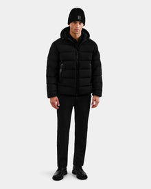 A man faces forward wearing the Rudsak DEMIAN M - M123507 Black men’s down puffer jacket with a hood, made from sustainable recycled fabric, paired with black pants and shoes against a plain light gray background.