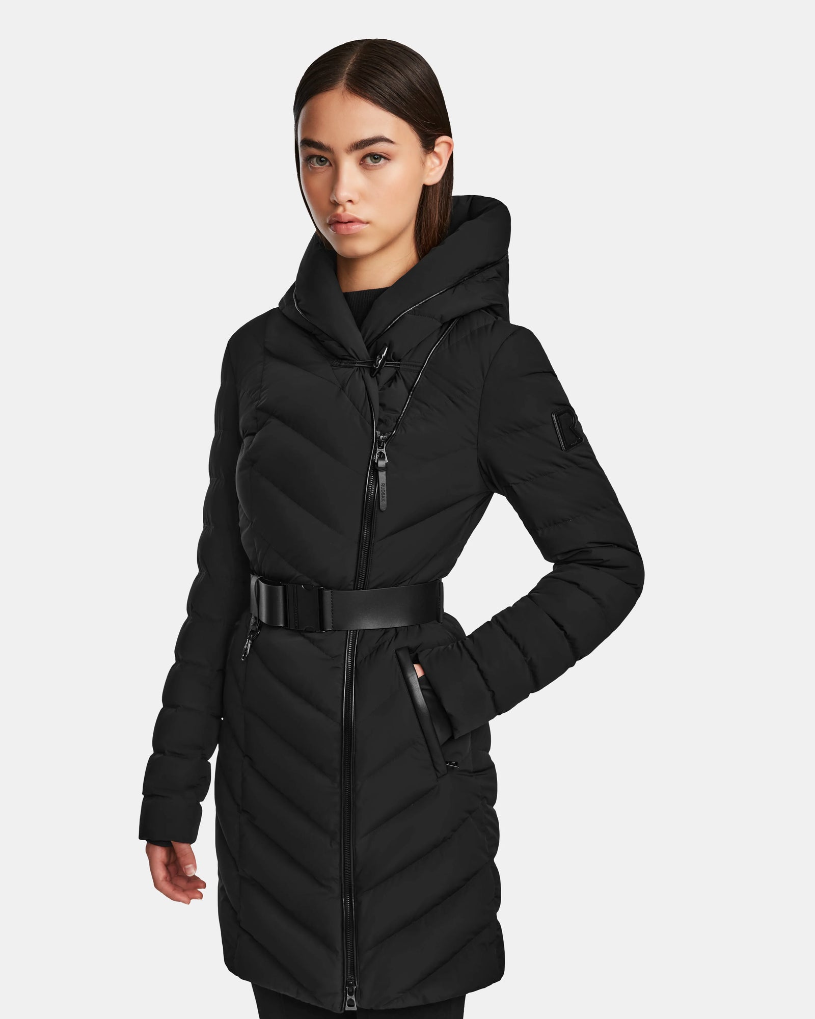 A woman models the Rudsak DITA - L124556 Black knee-length puffer with a belted waist, hood, diagonal quilting, zippered front, and RDS-certified down fill against a light background.