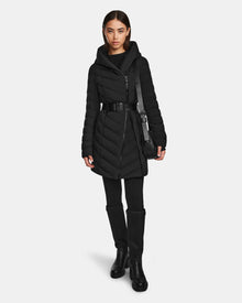 A woman poses in the Rudsak DITA - L124556 Black knee-length puffer jacket with a belted waist, paired with black pants and boots. She has straight brown hair, a neutral expression, and carries a black bag against a plain background.
