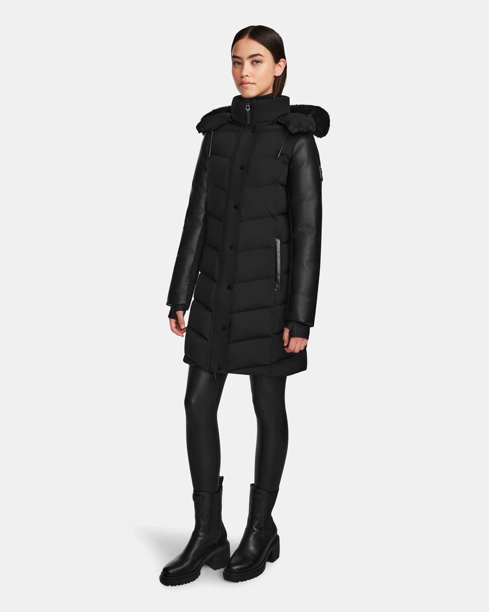 A woman faces forward, wearing the Rudsak ELSA - L123535 Black long quilted down puffer with high collar and hood, paired with black leggings and boots, against a plain white background.