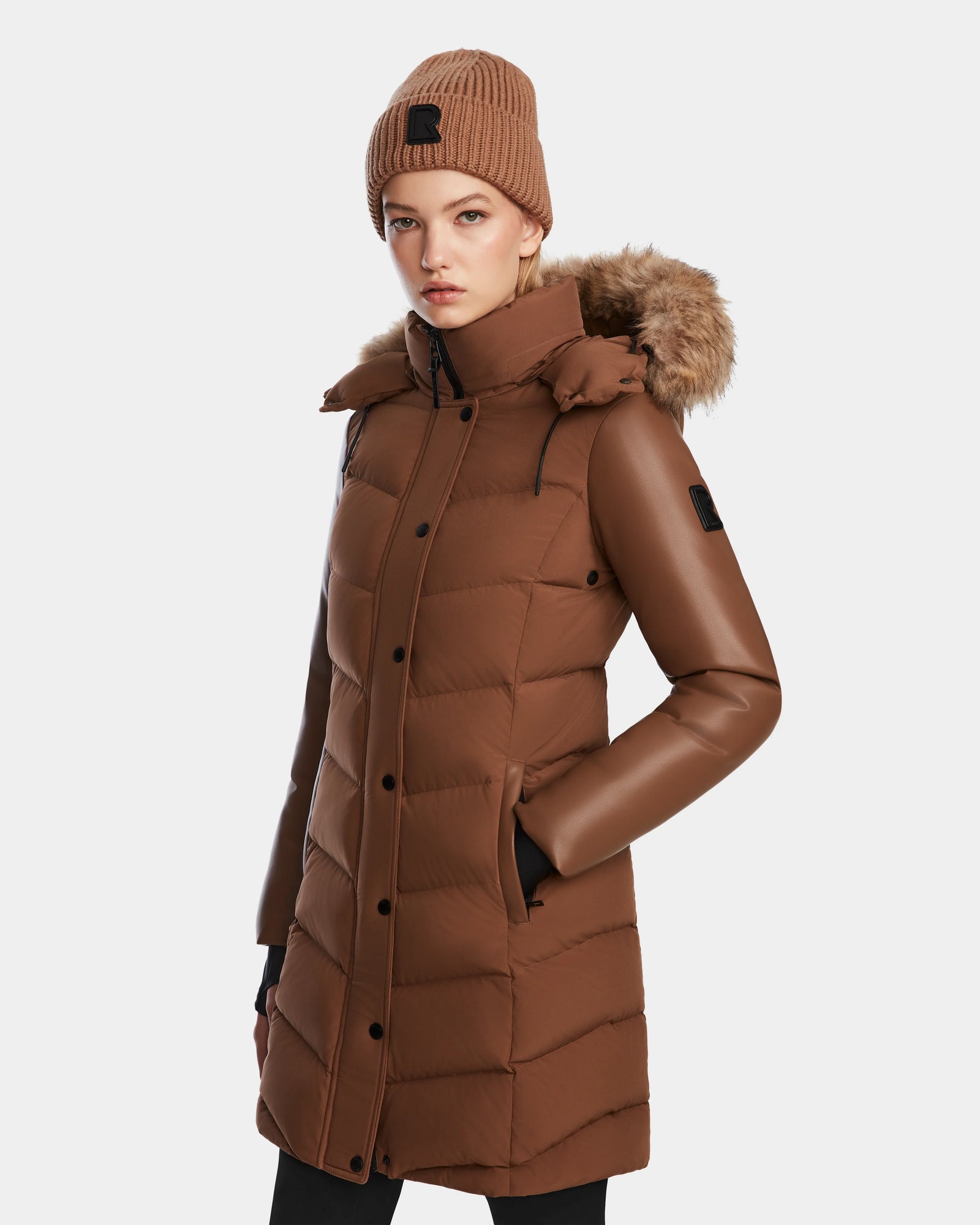 A woman wearing the Rudsak ELSA - L123535 Taupe knee-length quilted down puffer coat with a fur-lined hood and matching brown knit beanie stands with her hands in her pockets, facing forward against a plain white background.