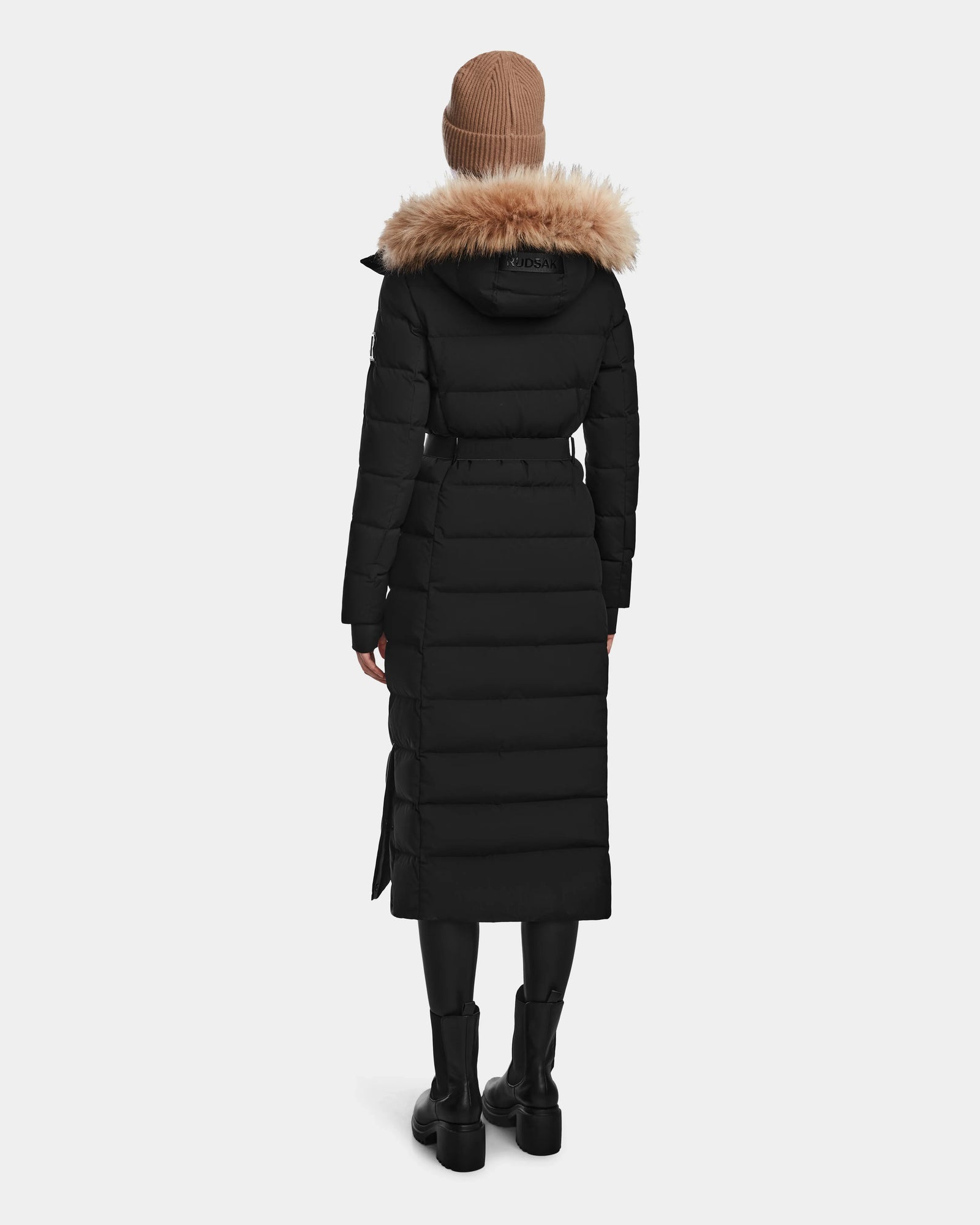 A person wearing the Rudsak ELSIE M - L123517 Black/Natural women’s maxi down puffer with a fur-trimmed hood, black boots, and a beige knit beanie stands facing away against a plain white background.