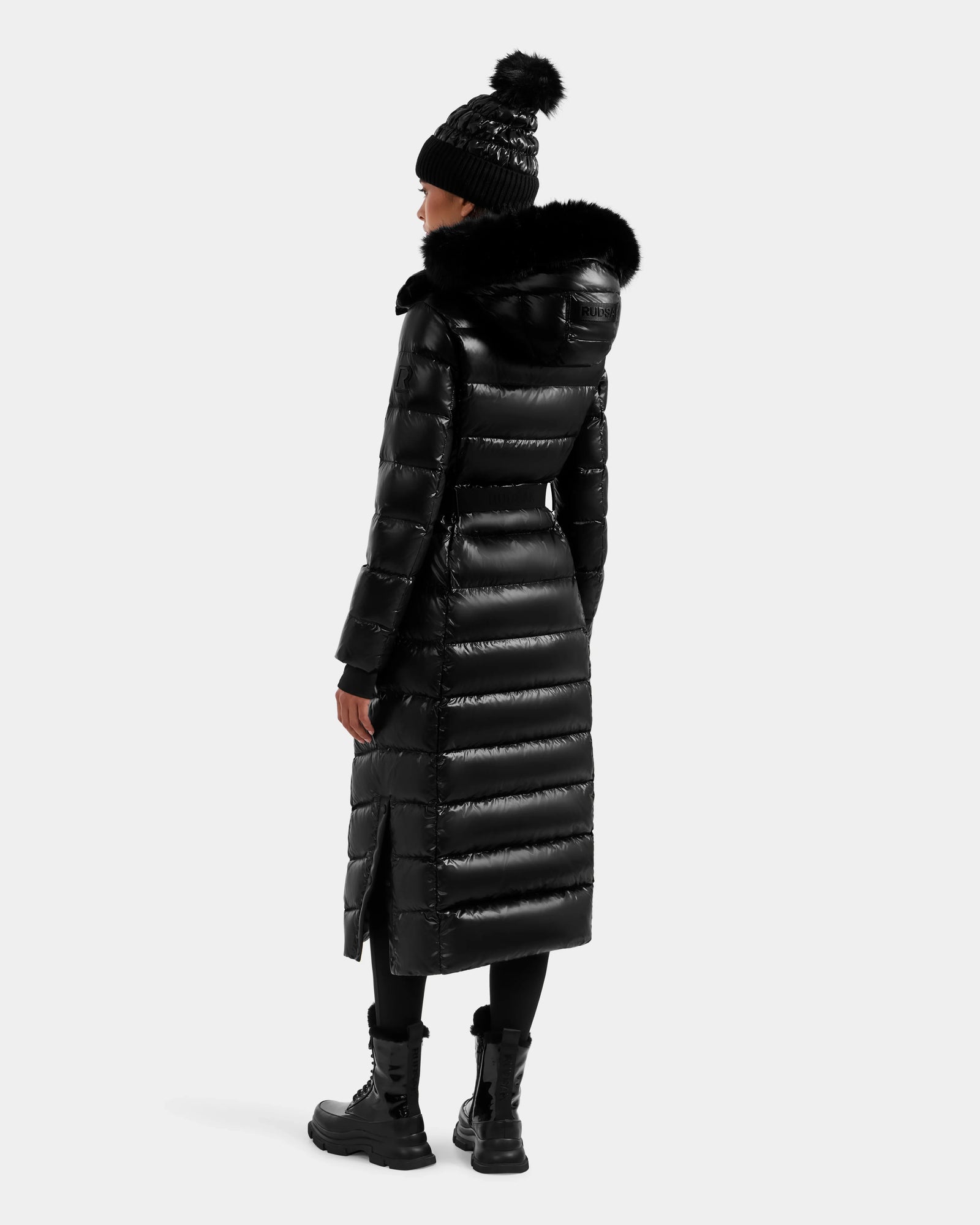 A person wearing the Rudsak ELSIE S - L124536 Black water-resistant women's maxi down puffer with a fur-lined hood, paired with black leggings, lace-up boots, and a pom-pom knit hat stands against a plain light background.