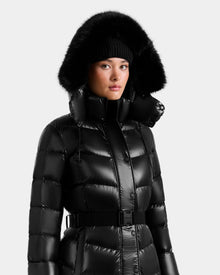 A person wears the Rudsak ELSIE S - L124536 Black women’s maxi puffer coat with a belted waist, high collar, and black fur-trimmed hood, paired with a black knit beanie against a plain, light background.