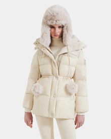 A woman wears the Rudsak EMBER M-M - L125524 Vanilla down puffer jacket with a fur-trimmed hood and RDS-certified down, paired with matching pom-poms, beige turtleneck sweater, and pants against a plain white background.