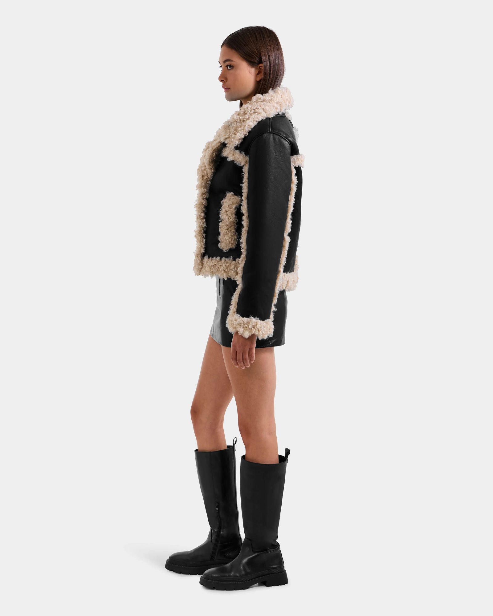 A woman stands in profile against a plain light background, wearing the Rudsak EMERY - L125548 Black vegan-leather jacket with beige faux shearling trim, paired with a black mini skirt and knee-high boots.