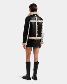 A woman with straight brown hair models the Rudsak EMERY - L125548 Black women's faux shearling jacket with light tan trim, a black mini skirt, and knee-high boots, standing against a white background facing away from the camera.