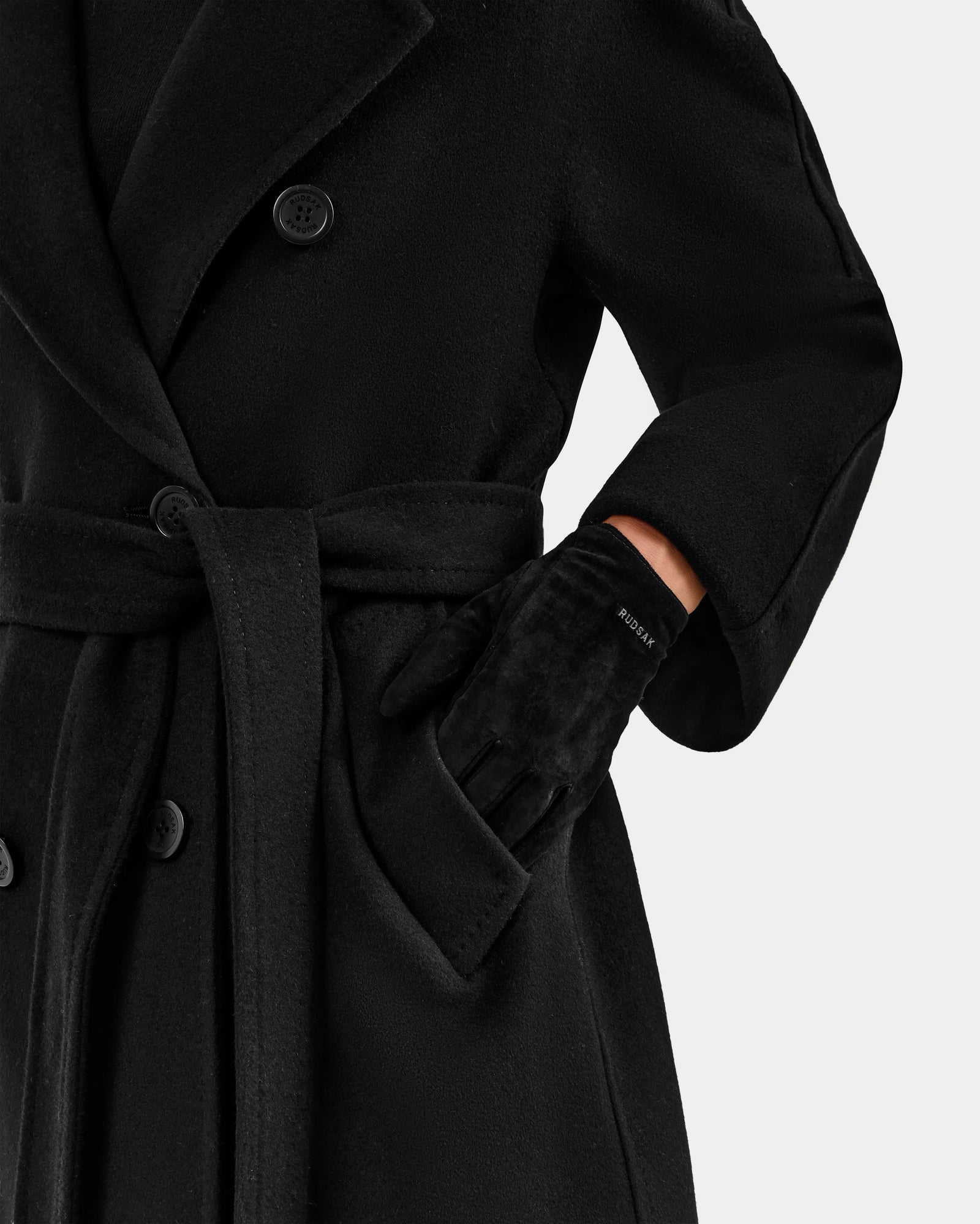 A person wears the Rudsak FLORA - L425542 Black double-breasted coat with a belt and winter gloves. Their gloved right fist is tucked in a pocket, with the photo capturing only the torso and arm, not the face.