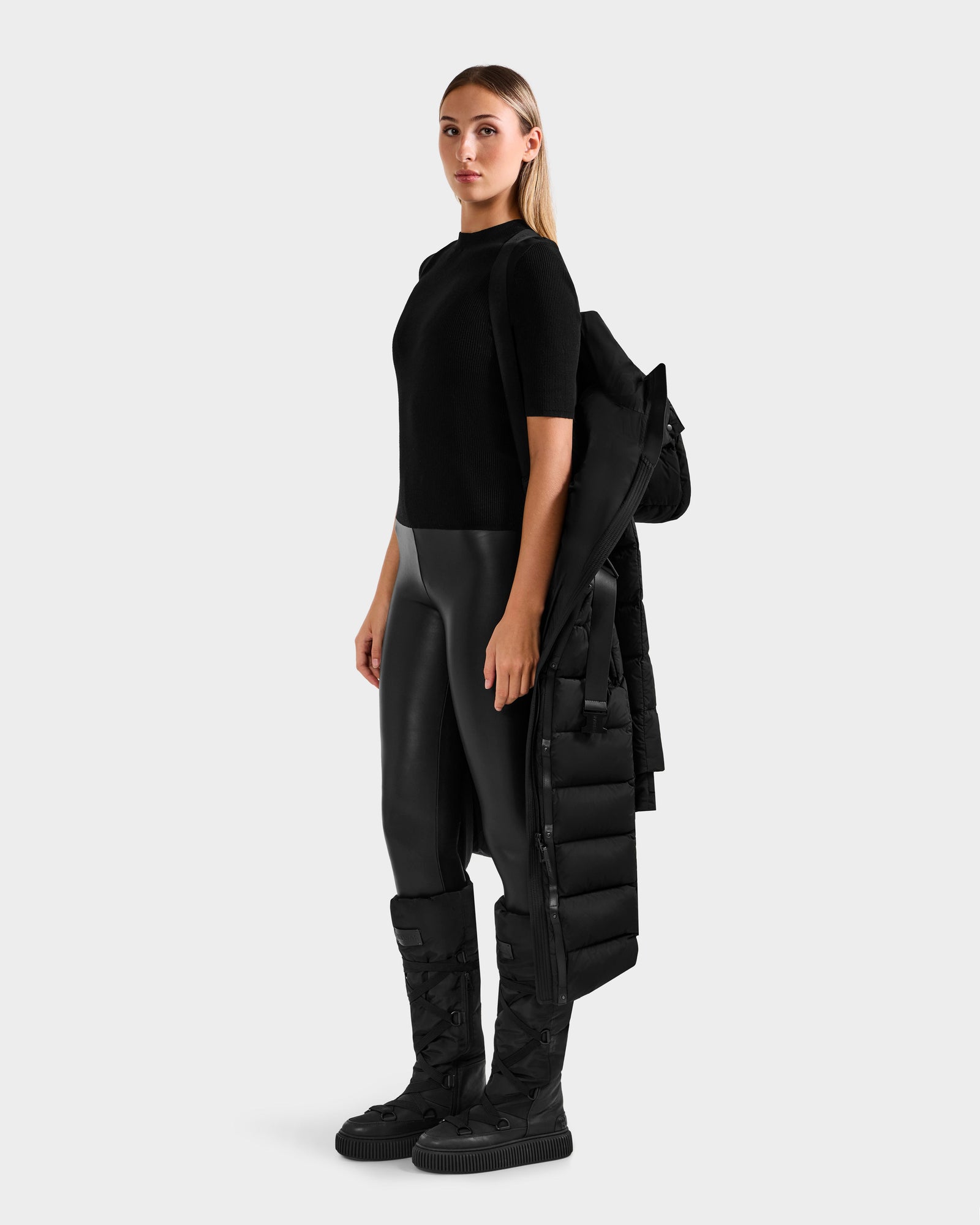 A woman stands facing forward, wearing the Rudsak FRIDA M-L - L125608 Black quilted down coat draped over one arm against a plain light gray background.