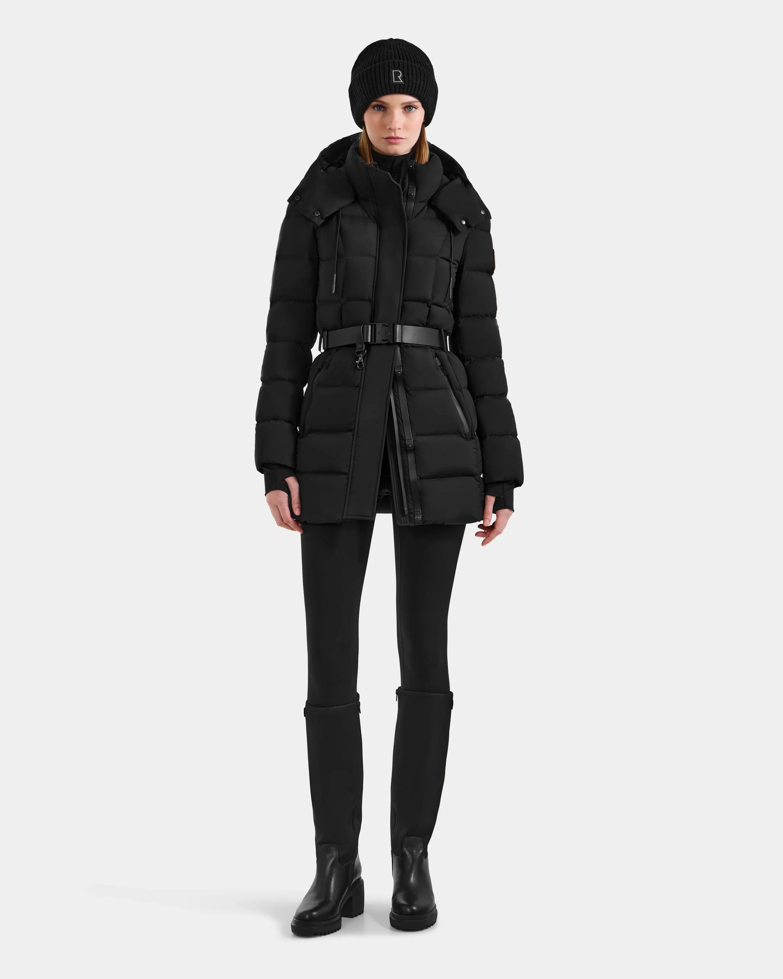 A person stands facing forward wearing the Rudsak FRIDA M-M - L125607 Black down puffer with a belt and detachable hood, styled with slim pants, knee-high boots, and a knit beanie hat against a plain white background.